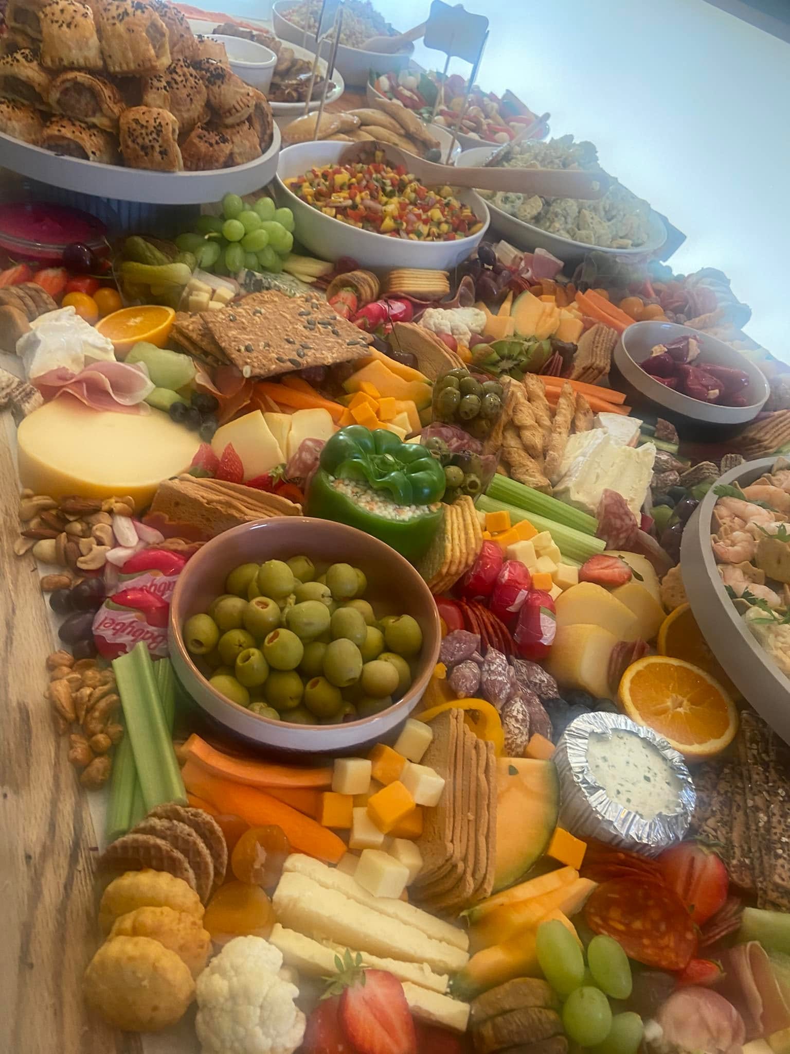 An abundant grazing table of sausage rolls, cheeses, charcuterie, olives, a stuffed pepper, crackers and fresh fruit